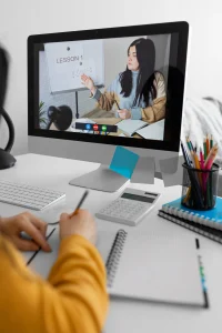 Student taking online math lesson from virtual tutor on desktop computer with study materials.