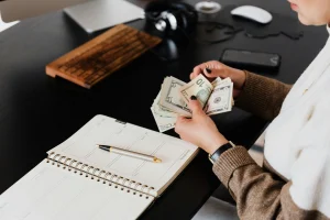 The image shows a person sitting at a desk, counting U.S. dollar bills, with a planner and pen in front of them. This scene represents earning money from home, potentially through remote or freelance work such as transcription, data entry, or budgeting freelance income.