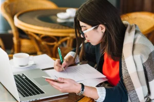Woman writing notes from laptop and phone while working on eBook or digital guide content creation remotely.