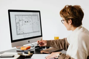 Architect working on building floor plan design on desktop computer while taking notes in modern office