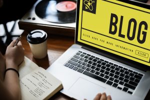 Person writing notes next to laptop displaying blog page with coffee and vinyl record on desk, ideal for content creation and blogging ideas.