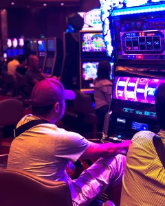 Person playing a slot machine in a brightly lit casino, surrounded by colorful lights and other gaming machines.