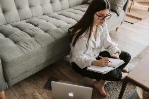 Young woman working from home, writing in notebook while sitting on floor with laptop, representing freelance or remote writing work.