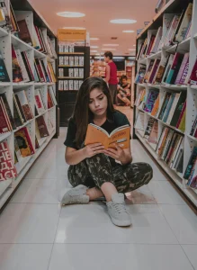 Young woman sitting on bookstore floor reading a book, surrounded by shelves filled with colorful books