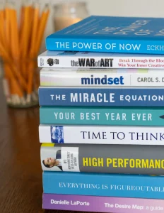 Stack of self-help books including "The Power of Now," "Mindset," and "The War of Art" on wooden desk with pencils in background