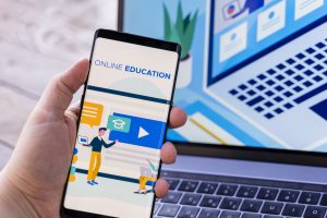 Hand holding smartphone displaying online education app with video lesson and graduation cap icon, laptop in background.