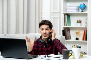 Man working from home, talking on phone while using laptop, wearing headphones, with books, globe, and office supplies in background.
