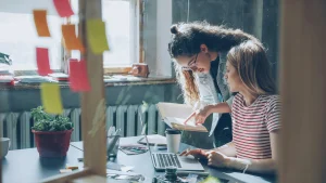 Two young women collaborating on a project with laptop and book at a creative workspace with sticky notes and plants