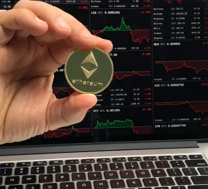 Hand holding Ethereum coin in front of laptop with cryptocurrency trading charts showing market fluctuations