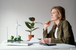 Woman thinking at desk with laptop, wind turbine model, solar panel, and plant, symbolizing sustainable energy and eco-friendly innovation