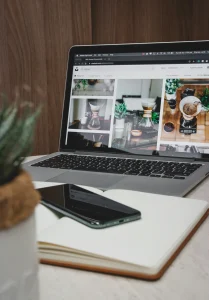Person browsing a website on a laptop, displaying images of coffee-related content, with notebooks and a plant in the foreground