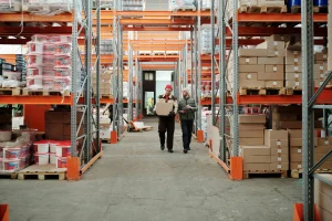 Workers with boxes in a warehouse with orange shelves and pallets.