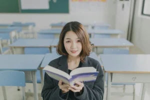 Student sitting in a classroom, holding an open book and smiling warmly at the camera.