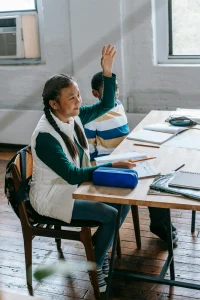 Two students sitting at a desk in a classroom, one raising their hand enthusiastically while the other looks on attentively.