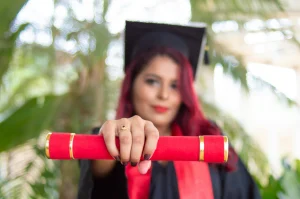 A graduate with red hair in a cap and gown proudly holds a red diploma with gold trim, standing against a blurred green foliage background.